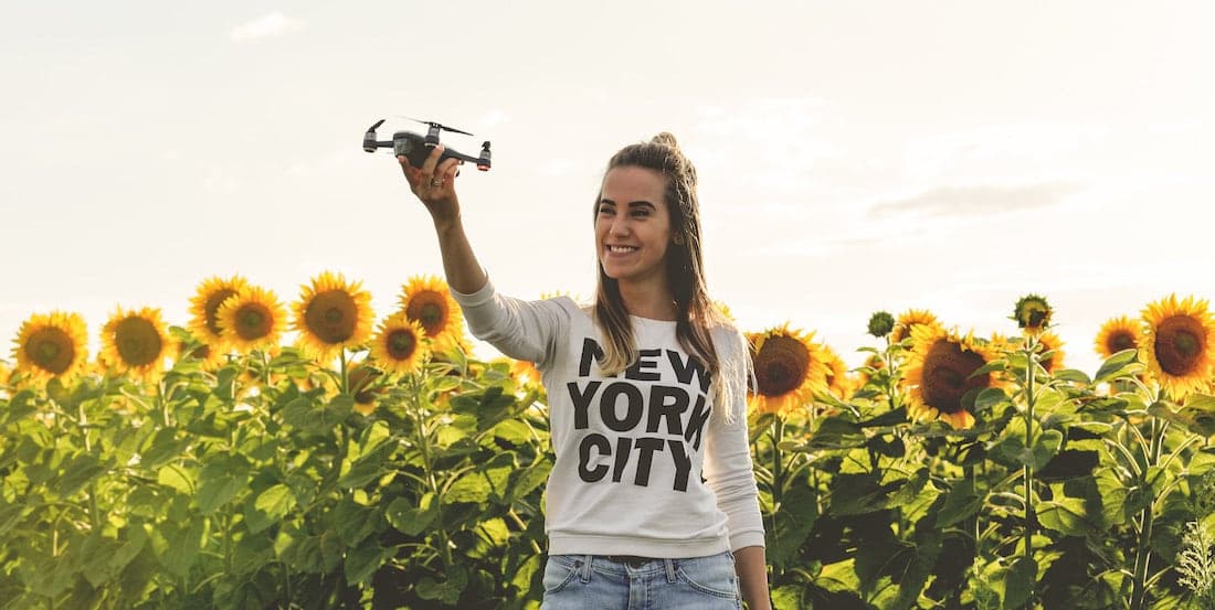 mujer joven con sudadera de nyc sujetando un dron con fondo de campo de girasoles y luz dorada de atardecer